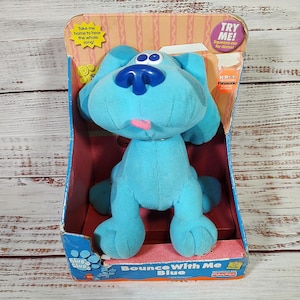 May include: A plush, light blue Blue's Clues toy dog in its original packaging. The toy has a large head, floppy ears, and a pink tongue. The packaging reads "Bounce With Me Blue" and includes the text "Try Me!"