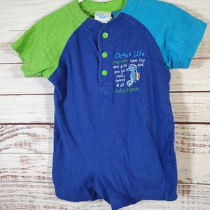 May include: A blue, green, and turquoise baby romper with short sleeves. The romper has a button-up front and features the text "Ocean Life" with a seahorse graphic. The sleeves are green and turquoise, and the romper is hanging on a white hanger.