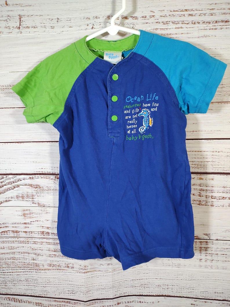 May include: A blue, green, and turquoise baby romper with short sleeves. The romper has a button-up front and features the text "Ocean Life" with a seahorse graphic. The sleeves are green and turquoise, and the romper is hanging on a white hanger.