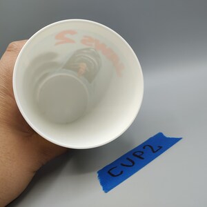 May include: A white cup with orange text inside, held by a hand. The text is partially visible. A blue piece of tape with "CUP 2" written on it is in the lower right corner.
