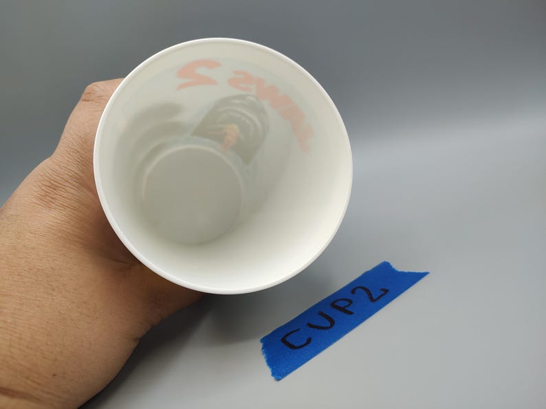 May include: A white cup with orange text inside, held by a hand. The text is partially visible. A blue piece of tape with "CUP 2" written on it is in the lower right corner.