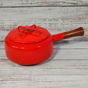 May include: A bright red saucepan with a matching lid and a wooden handle. The lid has a cross-shaped handle for easy lifting. The pan is designed for stovetop cooking and is a vibrant addition to any kitchen.