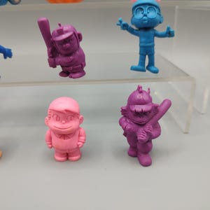 K&ouml;nnte beinhalten: Eine Sammlung bunter, kleiner Figuren. Die Figuren sind in verschiedenen Farben gehalten, darunter Blau, Rosa, Orange und Lila. Einige Figuren halten Baseballschl&auml;ger, w&auml;hrend andere sich in verschiedenen Posen befinden. Die Figuren werden auf einem transparenten, mehrstufigen Regal ausgestellt.