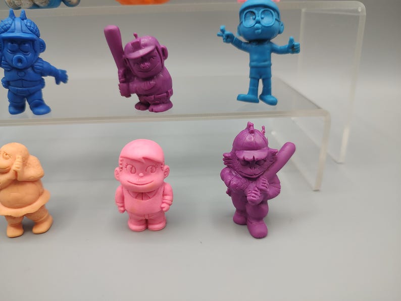 K&ouml;nnte beinhalten: Eine Sammlung bunter, kleiner Figuren. Die Figuren sind in verschiedenen Farben gehalten, darunter Blau, Rosa, Orange und Lila. Einige Figuren halten Baseballschl&auml;ger, w&auml;hrend andere sich in verschiedenen Posen befinden. Die Figuren werden auf einem transparenten, mehrstufigen Regal ausgestellt.