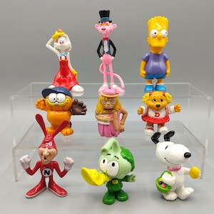May include: A collection of colorful cartoon character figurines, including Garfield, Bart Simpson, Snoopy, and the Pink Panther. The figures are made of plastic and are displayed on a clear shelf against a neutral background.