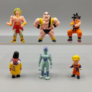 May include: A collection of six colorful Dragon Ball Z action figures. The figures depict various characters in dynamic poses, with vibrant colors and detailed designs. The figures are made of plastic and are displayed on a gray surface.