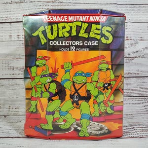 May include: A vintage Teenage Mutant Ninja Turtles collectors case. The case is rectangular with a red base and a blue top. The front features the words "Turtles Collectors Case Holds 12 Figures" and an illustration of the four turtles.
