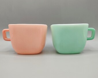 May include: Two vintage square-shaped coffee cups. One is a soft peach color, and the other is a light green. Both cups have a handle and a rounded base. The cups are made of a solid, opaque material.