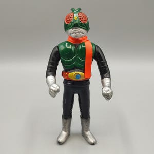 May include: A vintage action figure of a masked superhero. The figure has a green helmet with red eyes, an orange scarf, and a black and green torso. The figure is wearing black pants, silver boots, and silver gloves.