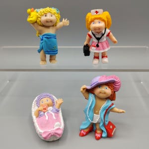 May include: Four Cabbage Patch Kids figurines. One is wrapped in a blue towel, another is dressed as a nurse, a third is in a pink bassinet, and the last is wearing a hat, blue shawl, and holding a red purse.