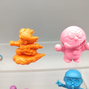 K&ouml;nnte beinhalten: Eine Sammlung farbenfroher Zeichentrickfiguren. Die Figuren umfassen eine blaue katzenartige Figur, eine orangefarbene Figur auf einer Wolke, eine rosa Figur mit einer Schleife und eine blaue Figur mit Brille. Dies sind wahrscheinlich Sammlerst&uuml;cke.