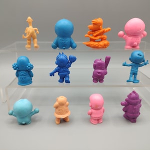 K&ouml;nnte beinhalten: Eine Sammlung farbenfroher Miniaturfiguren, darunter eine blaue Figur mit Baseballschl&auml;ger, eine rosa Figur mit erhobenen Armen und eine lila Figur mit Hut. Die Figuren werden auf einem transparenten, mehrstufigen Regal ausgestellt.