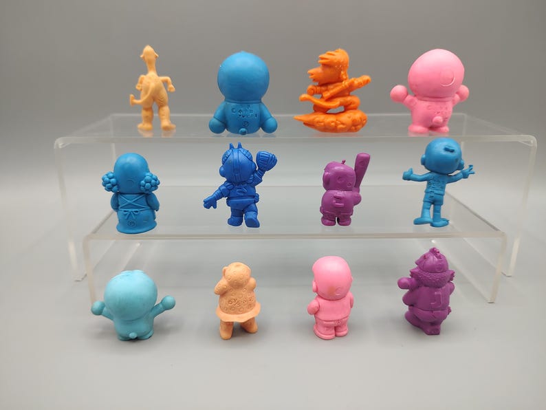 K&ouml;nnte beinhalten: Eine Sammlung farbenfroher Miniaturfiguren, darunter eine blaue Figur mit Baseballschl&auml;ger, eine rosa Figur mit erhobenen Armen und eine lila Figur mit Hut. Die Figuren werden auf einem transparenten, mehrstufigen Regal ausgestellt.