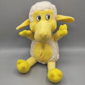 May include: A plush toy shaped like a sheep with a yellow face, yellow limbs, and a yellow belly. The sheep has a cream-colored, textured fleece body and head. It features large white eyes with black pupils and black eyelashes, and two long, floppy yellow ears.