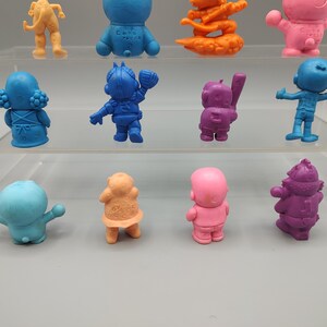 K&ouml;nnte beinhalten: Eine Sammlung farbenfroher, kleiner Figuren in verschiedenen Posen. Die Figuren sind in Blau-, Orange-, Rosa- und Lilat&ouml;nen gehalten. Einige Figuren haben Baseballschl&auml;ger, andere Helme. Die Figuren werden auf einem transparenten, mehrstufigen Regal ausgestellt.