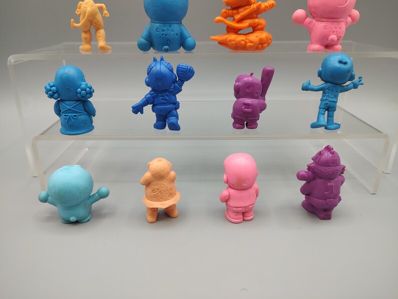 K&ouml;nnte beinhalten: Eine Sammlung farbenfroher, kleiner Figuren in verschiedenen Posen. Die Figuren sind in Blau-, Orange-, Rosa- und Lilat&ouml;nen gehalten. Einige Figuren haben Baseballschl&auml;ger, andere Helme. Die Figuren werden auf einem transparenten, mehrstufigen Regal ausgestellt.