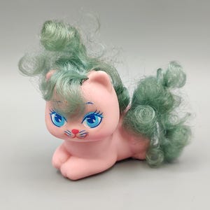 May include: A pink cat figurine with blue eyes and teal hair. The cat is lying down with its paws extended. The cat has a small pink nose and whiskers. The teal hair is styled in curls and a fluffy tail.