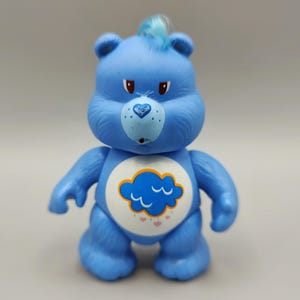 May include: A blue Care Bear figurine with a cloud and heart belly badge. The bear has a tuft of blue hair, a heart-shaped nose, and a grumpy expression. The figurine is standing upright.