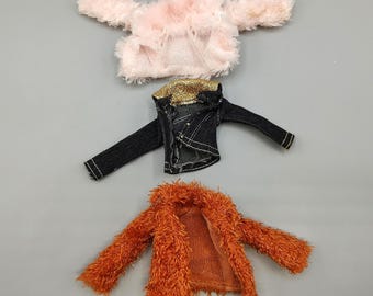 May include: Three doll-sized jackets in a top-down view. The jackets include a pink fuzzy jacket, a black denim jacket with gold accents, and an orange fuzzy jacket. The jackets are displayed on a gray surface.