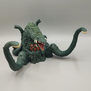 May include: A detailed, dark green monster figure with multiple tentacles and a textured body. The creature has a menacing open mouth with white teeth and a red interior. The figure's head and back are a lighter green, with a rough, scaly texture.