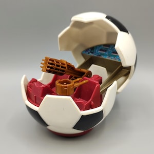 May include: A toy soccer ball, partially open, revealing a red interior with a brown gun-like structure and a smaller brown turret. The ball is white with black pentagons, and the interior has a blue circuit board.