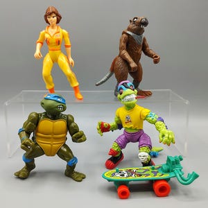 May include: A collection of Teenage Mutant Ninja Turtles action figures. The figures include April O'Neil in a yellow jumpsuit, a brown rat, a green turtle with a blue mask, and a lizard-like figure with a skateboard.