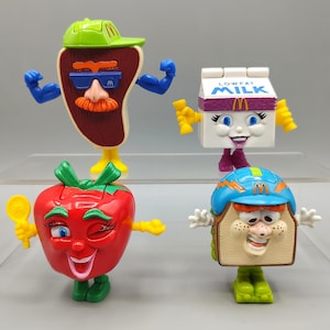 May include: Four McDonald's Happy Meal toys are displayed. They include a steak with a green visor, a carton of low-fat milk, a red tomato, and a sandwich, all with animated features and McDonald's branding.