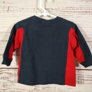 May include: A navy blue long-sleeved shirt with red side panels and sleeves. The shirt is hanging on a white hanger against a light wood background. The shirt is made of a soft material.