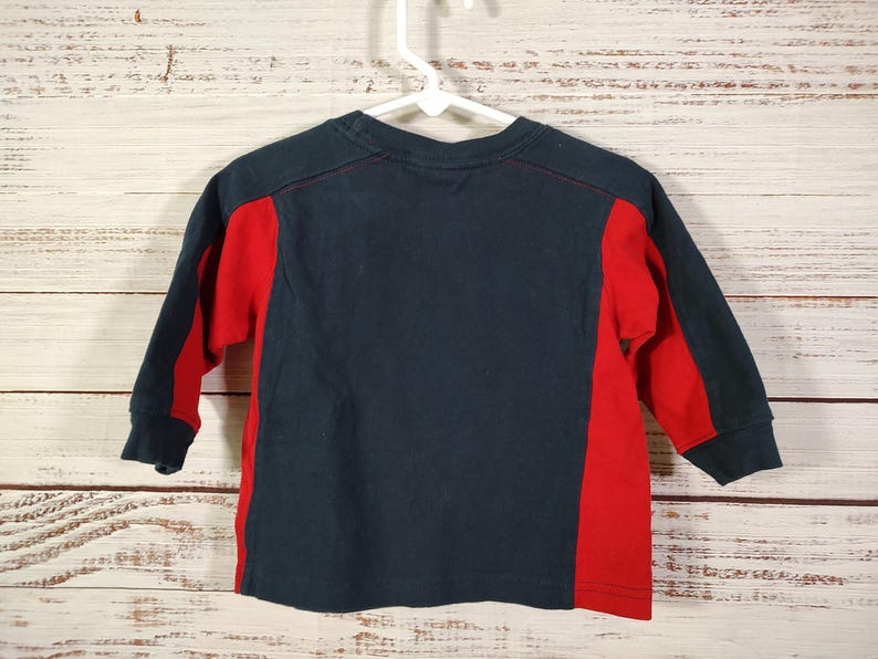 May include: A navy blue long-sleeved shirt with red side panels and sleeves. The shirt is hanging on a white hanger against a light wood background. The shirt is made of a soft material.