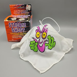 May include: A vintage Strobie Ghost toy with a white fabric ghost featuring a cartoon face with yellow eyes, pink mouth, and green hands. The box reads "Electronically Sound Activated Wild Dancing Motion".