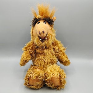 May include: A plush toy character with shaggy brown fur, a prominent snout, and dark brown eyebrows. The figure features a tuft of hair on top of its head, black button eyes, and an open mouth revealing white teeth against a dark interior.