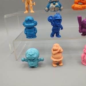 K&ouml;nnte beinhalten: Eine Sammlung farbenfroher, kleiner Figuren in verschiedenen Posen. Die Figuren sind in Blau-, Orange-, Rosa- und Lilat&ouml;nen gehalten. Einige stehen, andere befinden sich in Actionposen. Die Figuren werden auf einem transparenten, mehrstufigen Regal ausgestellt.