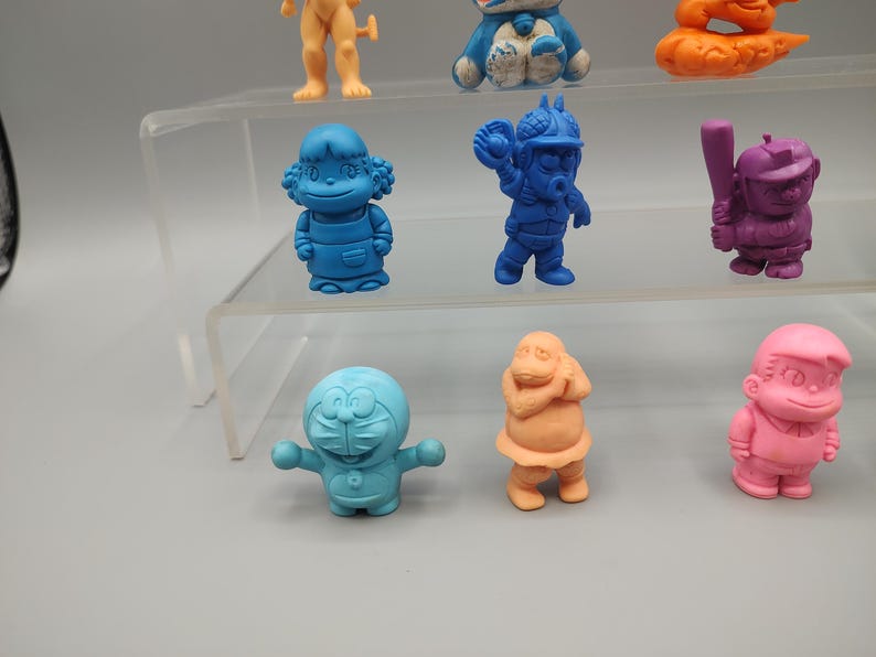 K&ouml;nnte beinhalten: Eine Sammlung farbenfroher, kleiner Figuren in verschiedenen Posen. Die Figuren sind in Blau-, Orange-, Rosa- und Lilat&ouml;nen gehalten. Einige stehen, andere befinden sich in Actionposen. Die Figuren werden auf einem transparenten, mehrstufigen Regal ausgestellt.