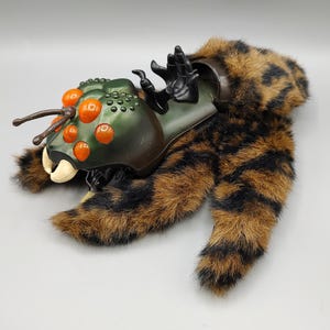 May include: A unique toy creature with a green and brown segmented body, orange accents, and furry brown and black legs. The toy has a detailed insect-like head with antennae and a set of black claws.