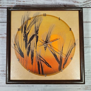 May include: Square wooden wall clock with a brown frame. The clock face features a wheat field scene with a setting sun in shades of orange and yellow. Black wheat stalks are silhouetted against the sunset. Silver clock hands.
