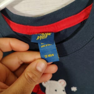 May include: Navy blue long-sleeved shirt featuring a white polar bear graphic with a red scarf. The shirt has a red trim around the neckline and a tag that says "12 Mos".