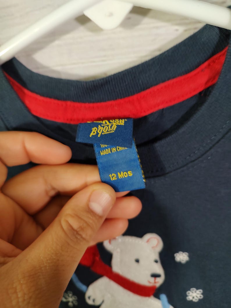May include: Navy blue long-sleeved shirt featuring a white polar bear graphic with a red scarf. The shirt has a red trim around the neckline and a tag that says "12 Mos".