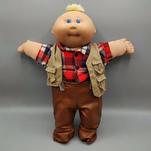 May include: A vintage doll with a bald head, blue eyes, and a tuft of blonde hair. The doll is wearing a red and black plaid shirt, a tan vest with pockets, and brown pants. The doll's arms are outstretched.