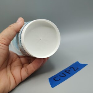 May include: A white plastic cup is held in a hand, with the bottom of the cup facing the camera. The cup has a rounded interior and a cylindrical exterior. A blue piece of tape with the text "CUP2" is in the lower right corner.