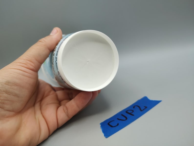 May include: A white plastic cup is held in a hand, with the bottom of the cup facing the camera. The cup has a rounded interior and a cylindrical exterior. A blue piece of tape with the text "CUP2" is in the lower right corner.