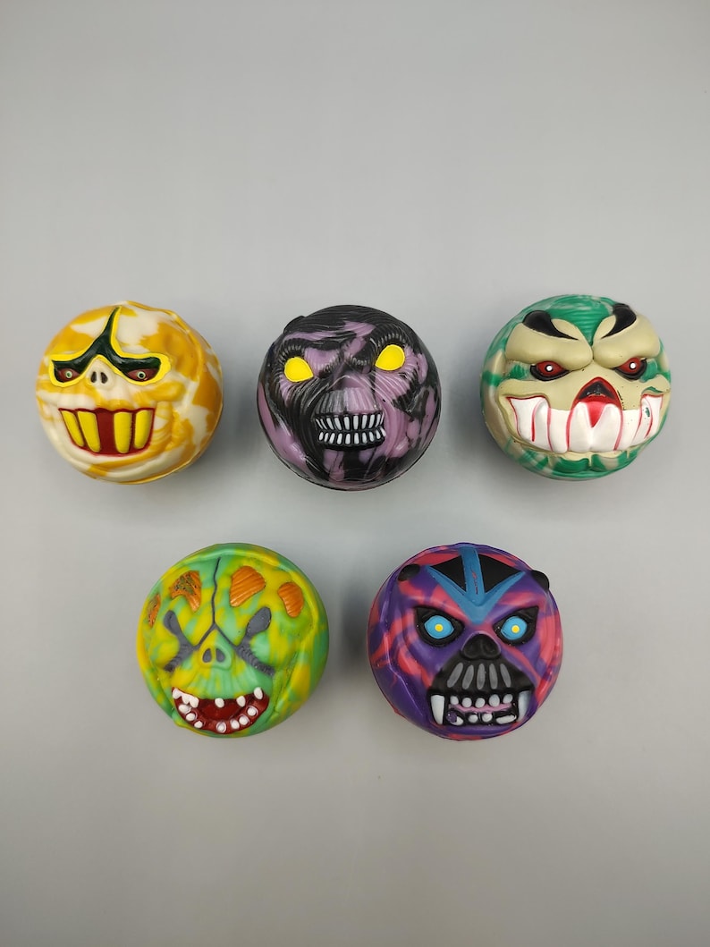 May include: Five colorful, round monster-faced toys. Each has a unique design with different colors, including yellow, purple, green, and blue. The faces feature exaggerated features like teeth and glowing eyes.