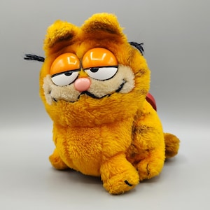 May include: A plush Garfield toy in a seated position. The orange cat has a cream-colored muzzle, a pink nose, and orange eyes. Black whiskers and a red heart-shaped tag are visible. The toy is made of soft, fuzzy material.