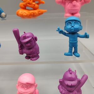 K&ouml;nnte beinhalten: Eine Sammlung farbenfroher Zeichentrickfiguren. Die Figuren sind in verschiedenen Posen und Farben, darunter Blau, Lila, Rosa und Orange. Einige Figuren halten Gegenst&auml;nde wie einen Baseballschl&auml;ger. Die Figuren werden auf transparenten Regalen ausgestellt.