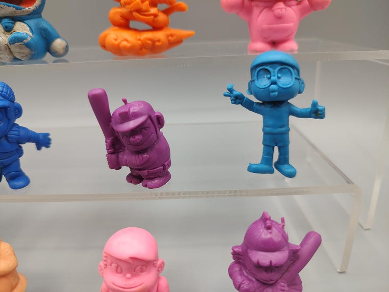 K&ouml;nnte beinhalten: Eine Sammlung farbenfroher Zeichentrickfiguren. Die Figuren sind in verschiedenen Posen und Farben, darunter Blau, Lila, Rosa und Orange. Einige Figuren halten Gegenst&auml;nde wie einen Baseballschl&auml;ger. Die Figuren werden auf transparenten Regalen ausgestellt.