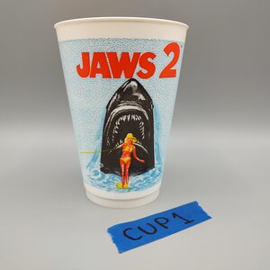 May include: A white paper cup featuring the movie title "JAWS 2" in red letters. The cup has a blue textured background with an illustration of a shark's open mouth, a woman in a swimsuit, and the text "CUP 1" on a blue tape.