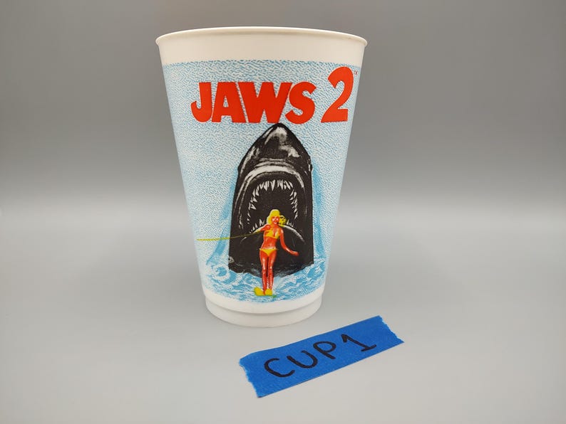 May include: A white paper cup featuring the movie title "JAWS 2" in red letters. The cup has a blue textured background with an illustration of a shark's open mouth, a woman in a swimsuit, and the text "CUP 1" on a blue tape.