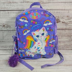 May include: A purple backpack featuring a cartoon cat with rainbow wings, hearts, and rainbows. The backpack has the text "Lisa Frank" and "Angel" printed on it. It includes a purple pom-pom and adjustable straps.