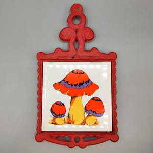 May include: A red cast iron trivet with a decorative scrollwork handle and frame, featuring a square white ceramic tile insert. The tile displays a colorful illustration of three mushrooms with orange caps, purple accents, and yellow stems.