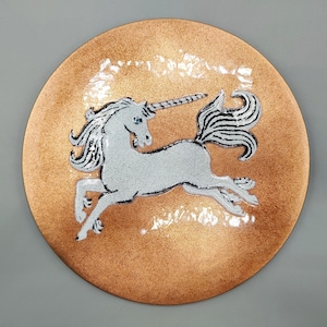 May include: A round, copper-colored decorative plate featuring a white unicorn with a black and white striped tail and horn. The unicorn is in a running pose, with its legs extended. The plate has a textured surface.