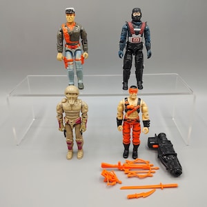 May include: Four action figures are displayed on a clear shelf. The figures are of various characters, each with unique outfits and accessories. One figure wears a military-style uniform, while another is clad in black with a red harness. Orange weapons are placed in front of the figures.